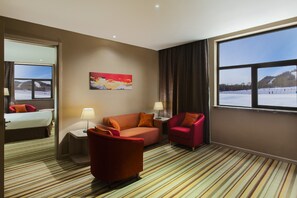 A hotel room with a large window, a sofa, a chair, a small table, and a painting on the wall.