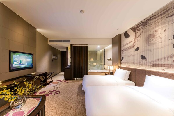 Habitación de hotel con televisor mostrando un mensaje de bienvenida, una cama, un escritorio con un jarrón de flores y un mural abstracto grande en la pared.