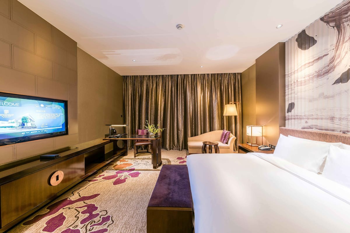 Ein Hotelzimmer mit einem großen Bett, einem Fernseher auf einem Schrank, einem Schreibtisch mit Stuhl, einer Couch und einem großen Fenster mit Vorhängen.