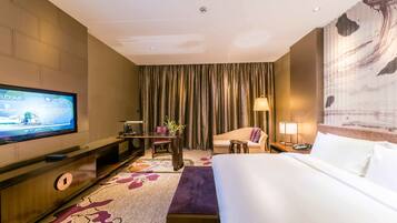 A hotel room with a large bed, a TV on a stand, a desk with a chair, a sofa, and a large window with curtains.