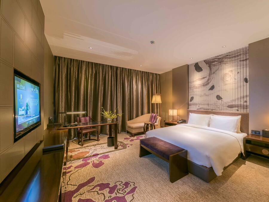 A hotel room with a large bed, a desk with a chair, a television, and a large window with curtains.