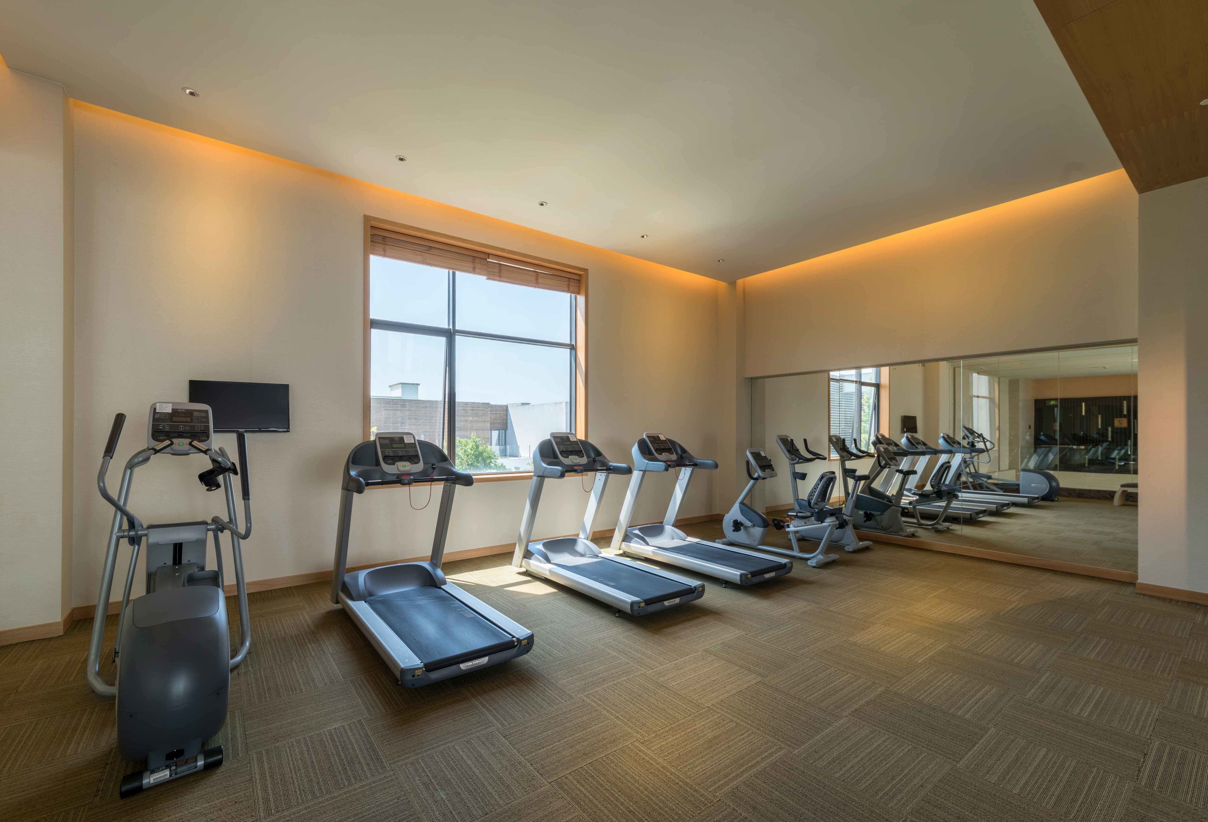 Un gimnasio con cintas de correr, elíptica y bicicleta estática, con ventanas amplias y un espejo en una pared.