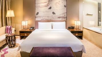 Habitación de hotel con una cama grande, mesitas de noche, un sofá, una pequeña mesa redonda y un baño con bañera y espejo.