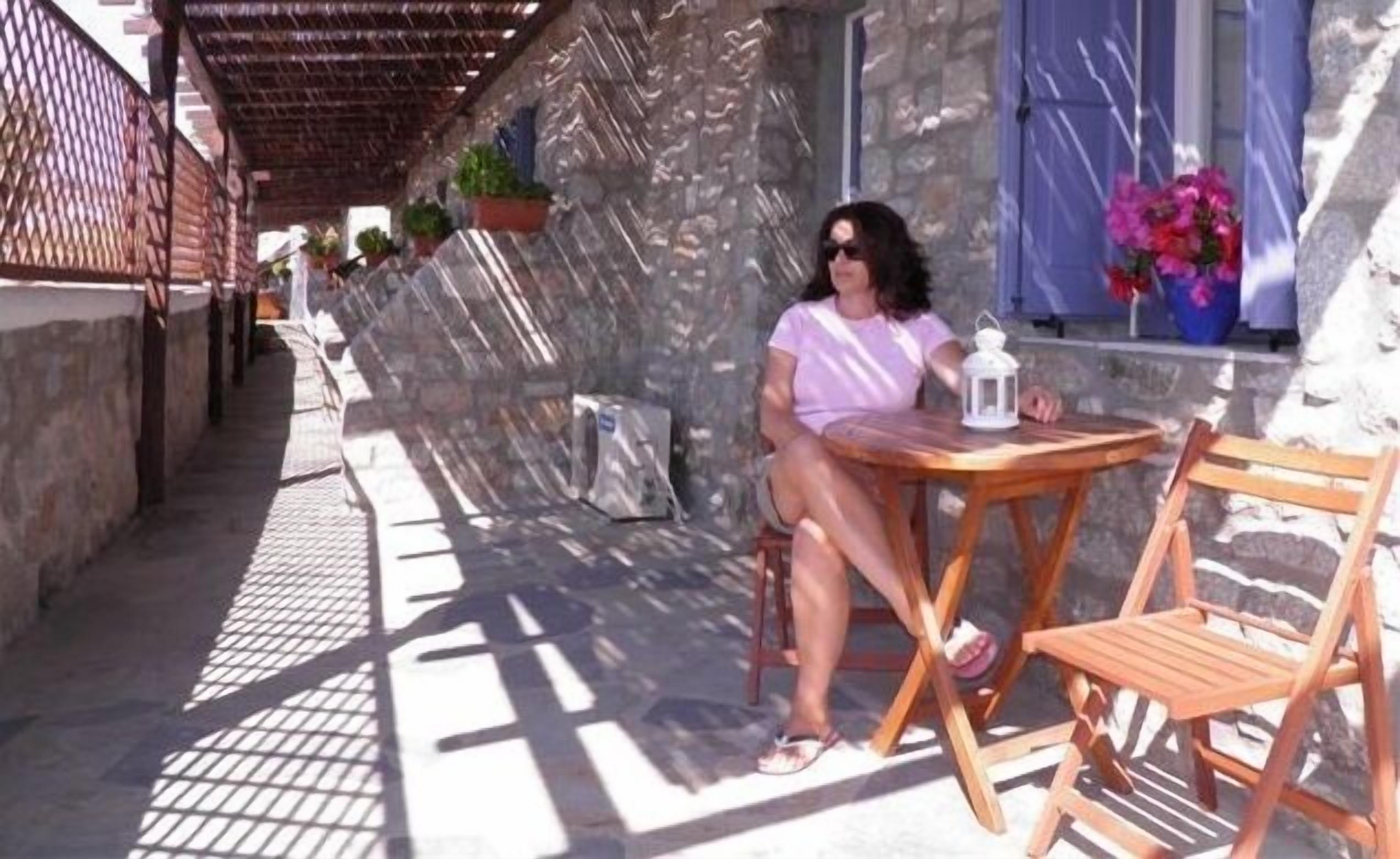A woman sitting at a wooden table with a lantern on it, in a shaded outdoor area with stone walls and potted plants.