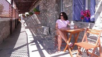 A woman sitting at a wooden table with a lantern on it, in a shaded outdoor area with stone walls and potted plants.