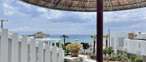A wooden table and chair under a thatched umbrella, overlooking a coastal area with a white fence and buildings.