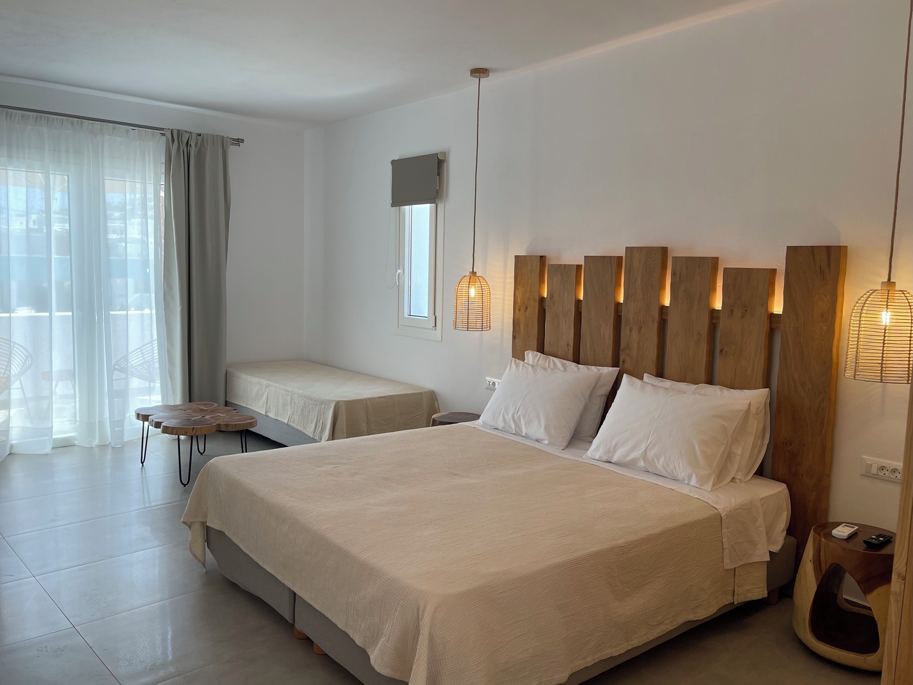 A modern hotel room with a large bed, a smaller bed, a wooden headboard, a bedside table, a lamp, and a view of the outdoors through sheer curtains.