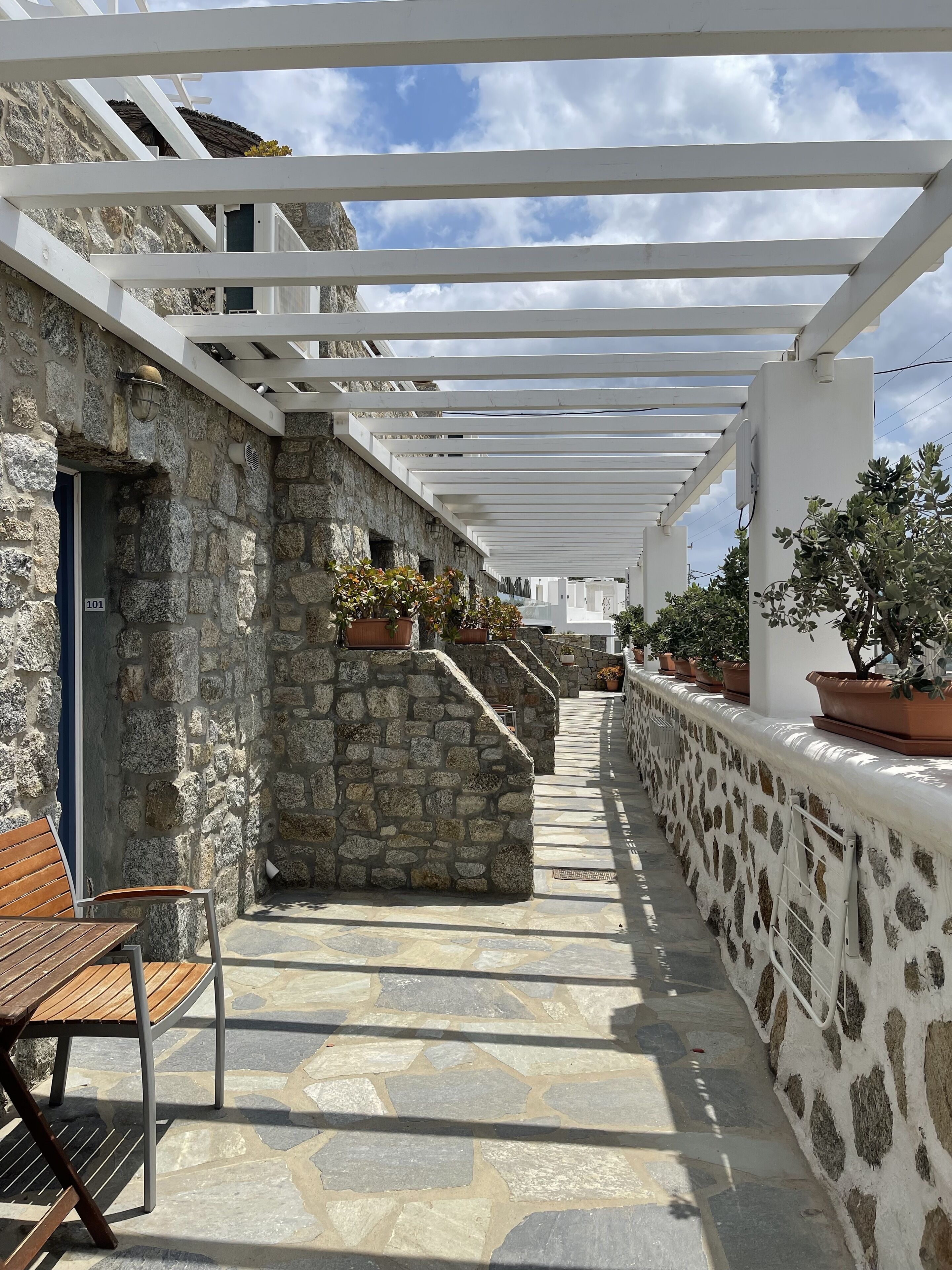 A stone-walled corridor with a wooden bench and potted plants, under a white pergola.