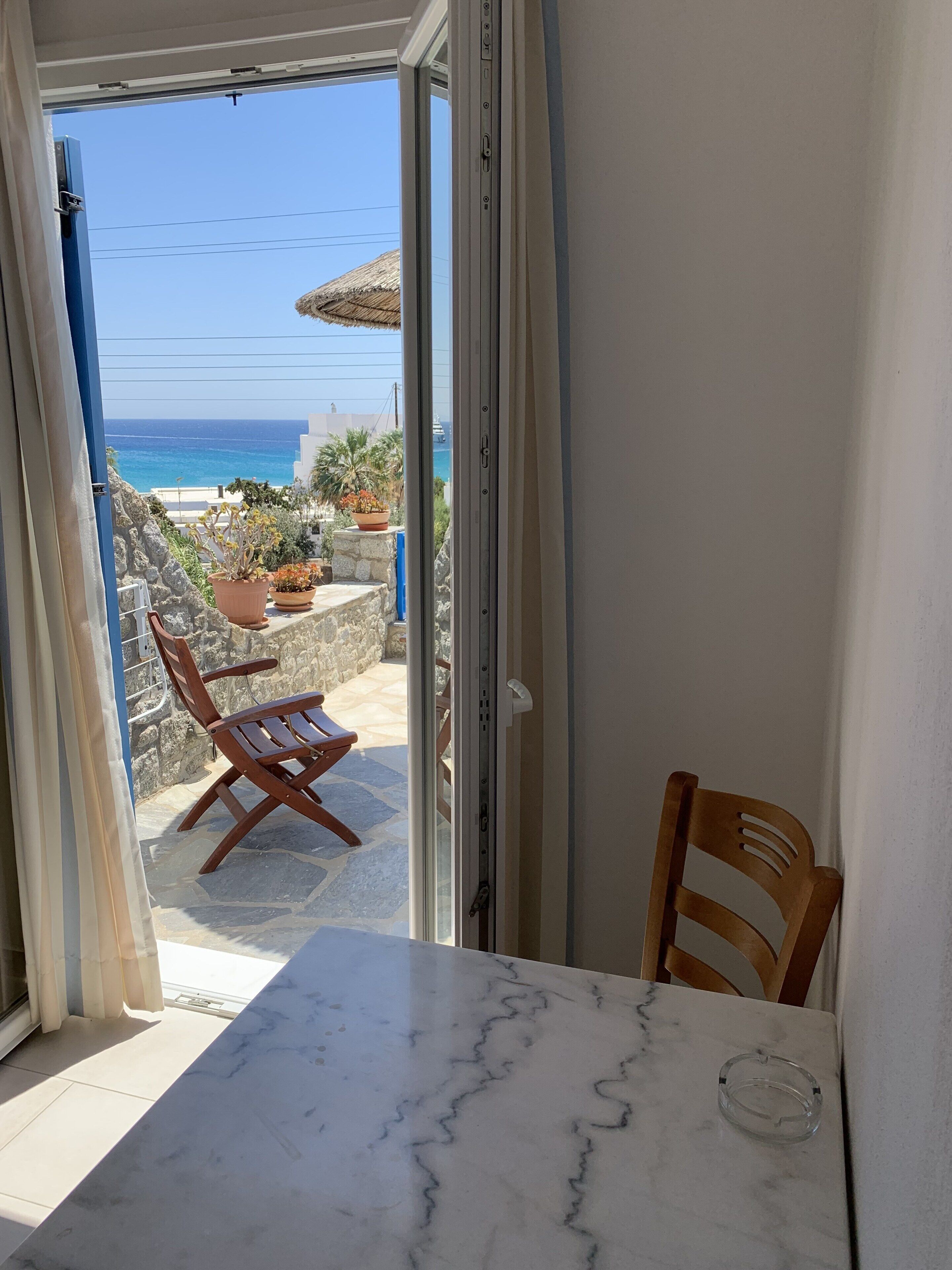 Una habitación con una mesa de mármol y una silla, vistas al mar y un patio con plantas y una mesa de madera fuera.