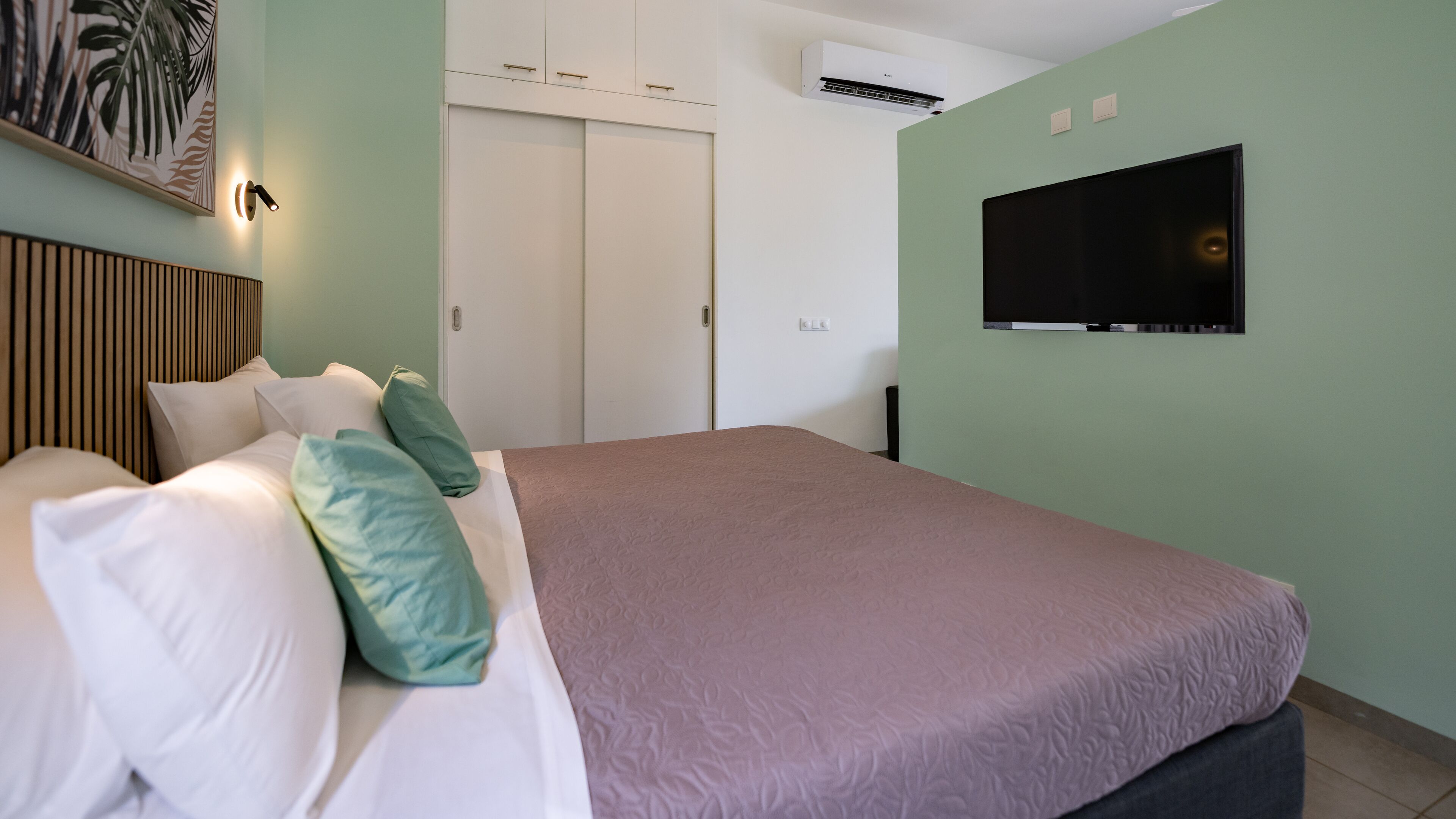 A bedroom with a bed, pillows, a TV, a wardrobe, and an air conditioning unit.