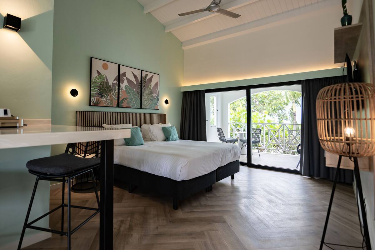 A modern bedroom with a large bed, a desk with a coffee maker, and a balcony with a view of greenery.