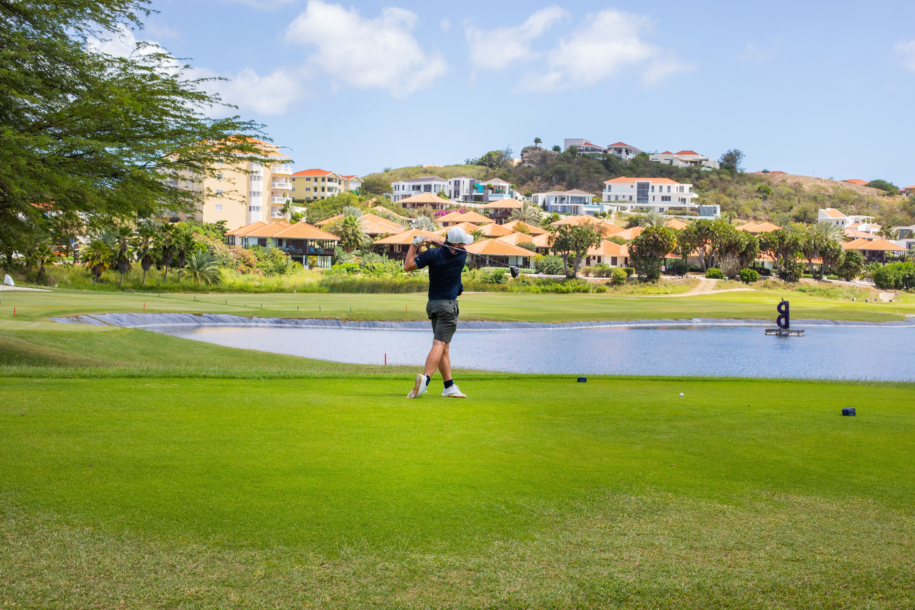 A golf course with a pond, a person in a blue shirt and shorts preparing to hit a golf ball, and a backdrop of residential buildings on a hill.