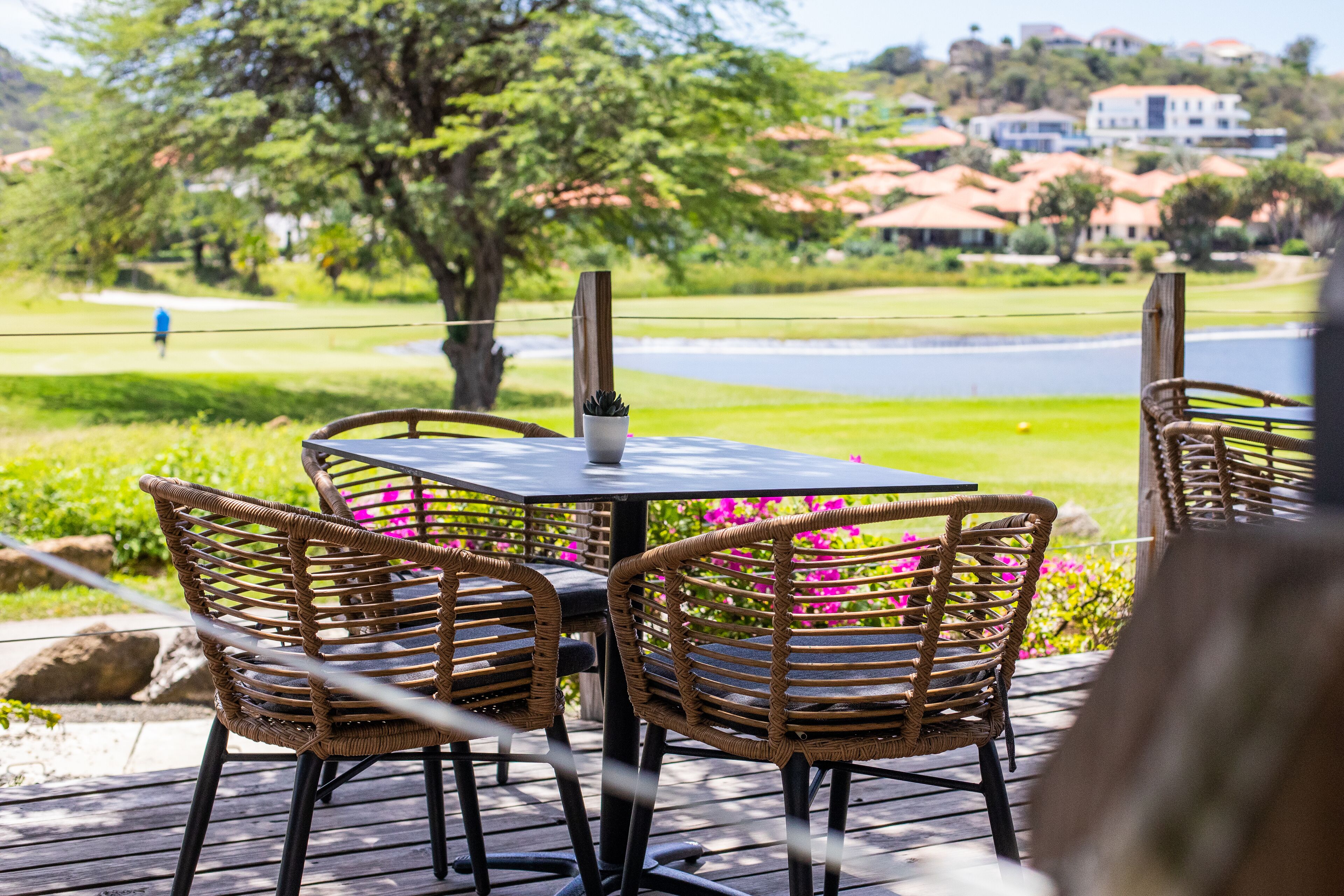 Un espacio de comedor al aire libre con sillas de mimbre y una mesa, con vista a un jardín ajardinado y una masa de agua.