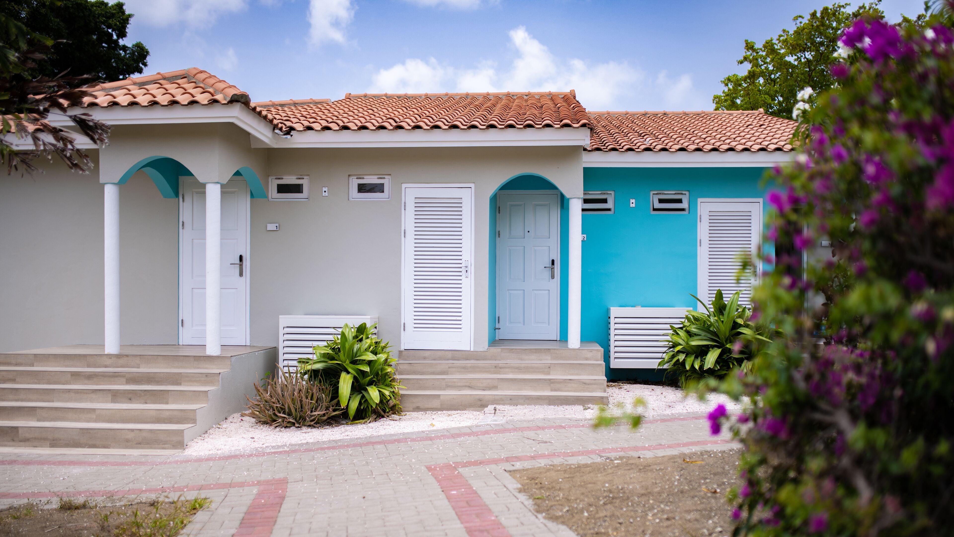A two-story building with a tiled roof, white and blue doors, and a small garden area with steps leading to the entrance.