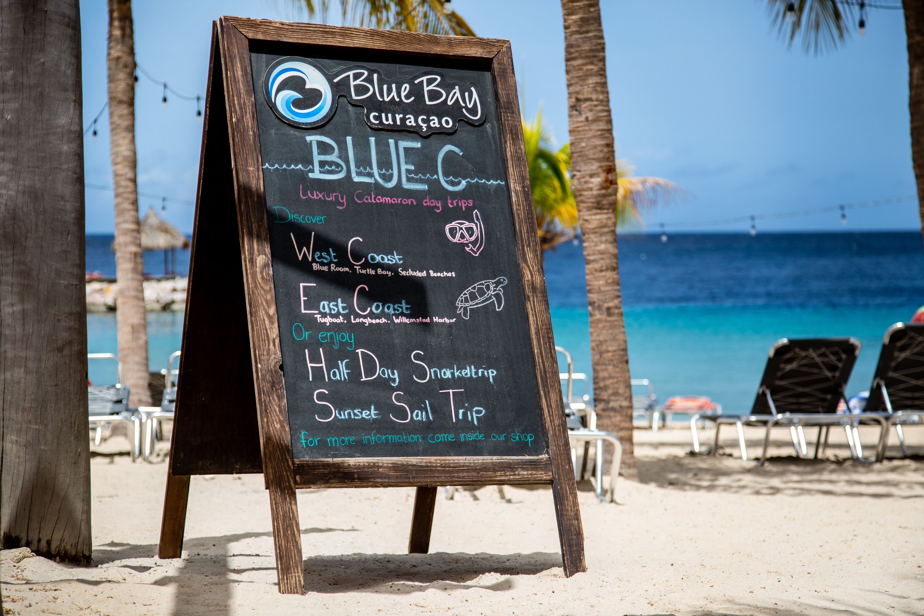 A beachside signboard advertising Blue Bay Curacao's luxury catamaran day trips, including West Coast and East Coast tours, with a view of the beach and ocean.