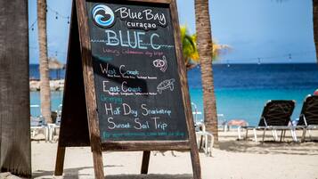 A beachside signboard advertising Blue Bay Curacao's luxury catamaran day trips, including West Coast and East Coast tours, with a view of the beach and ocean.