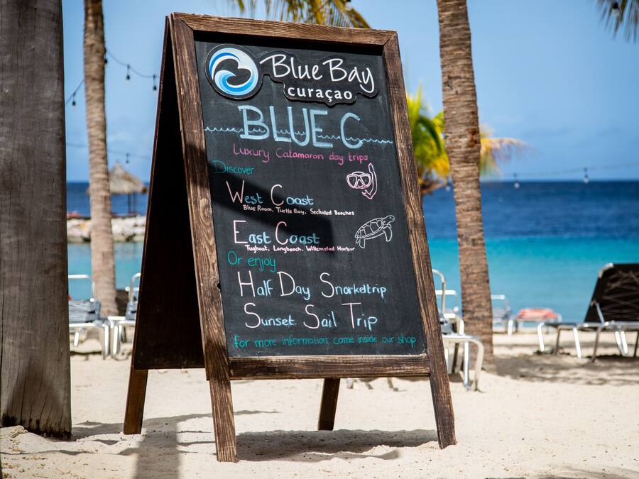 A beachside signboard advertising Blue Bay Curacao's luxury catamaran day trips, including West Coast and East Coast tours, with a view of the beach and ocean.
