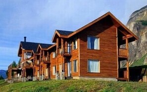 A wooden cabin with multiple balconies and large windows, situated on a grassy hill with a mountain in the background.