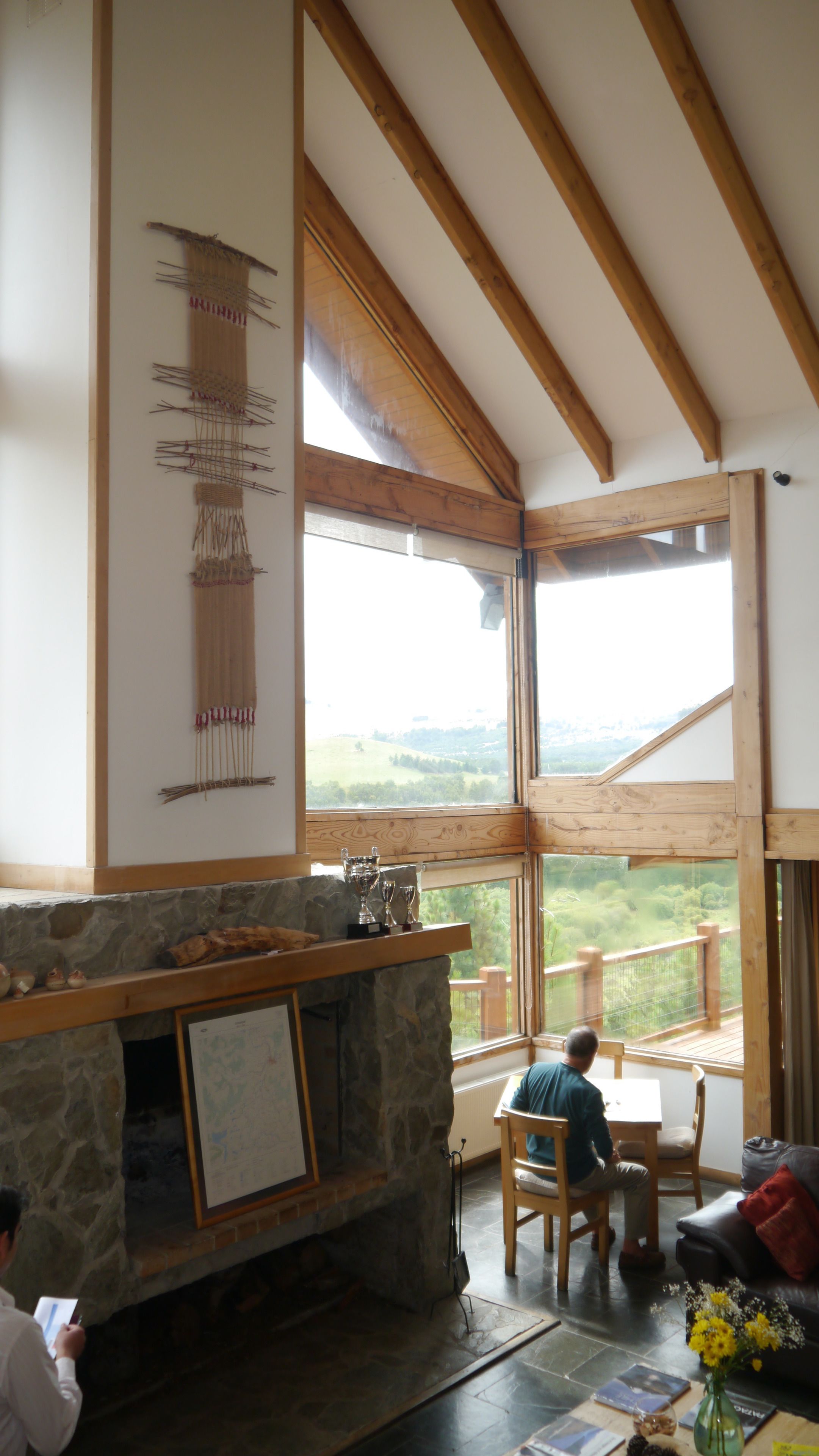A spacious room with a stone fireplace, wooden beams, and large windows offering a view of the countryside.