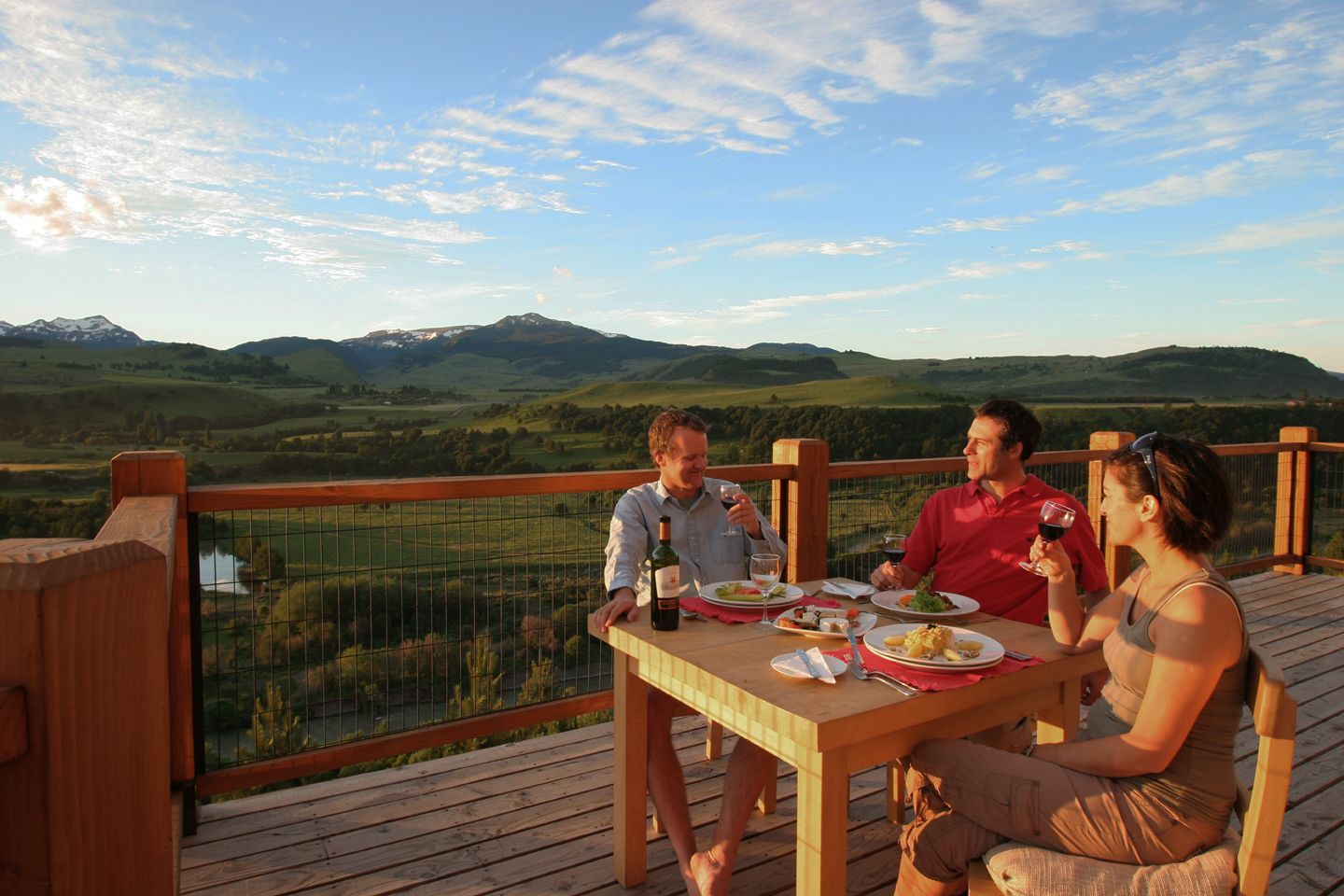 Una terraza de madera con una mesa dispuesta para tres personas, con vistas a un paisaje pintoresco con montañas y un lago.
