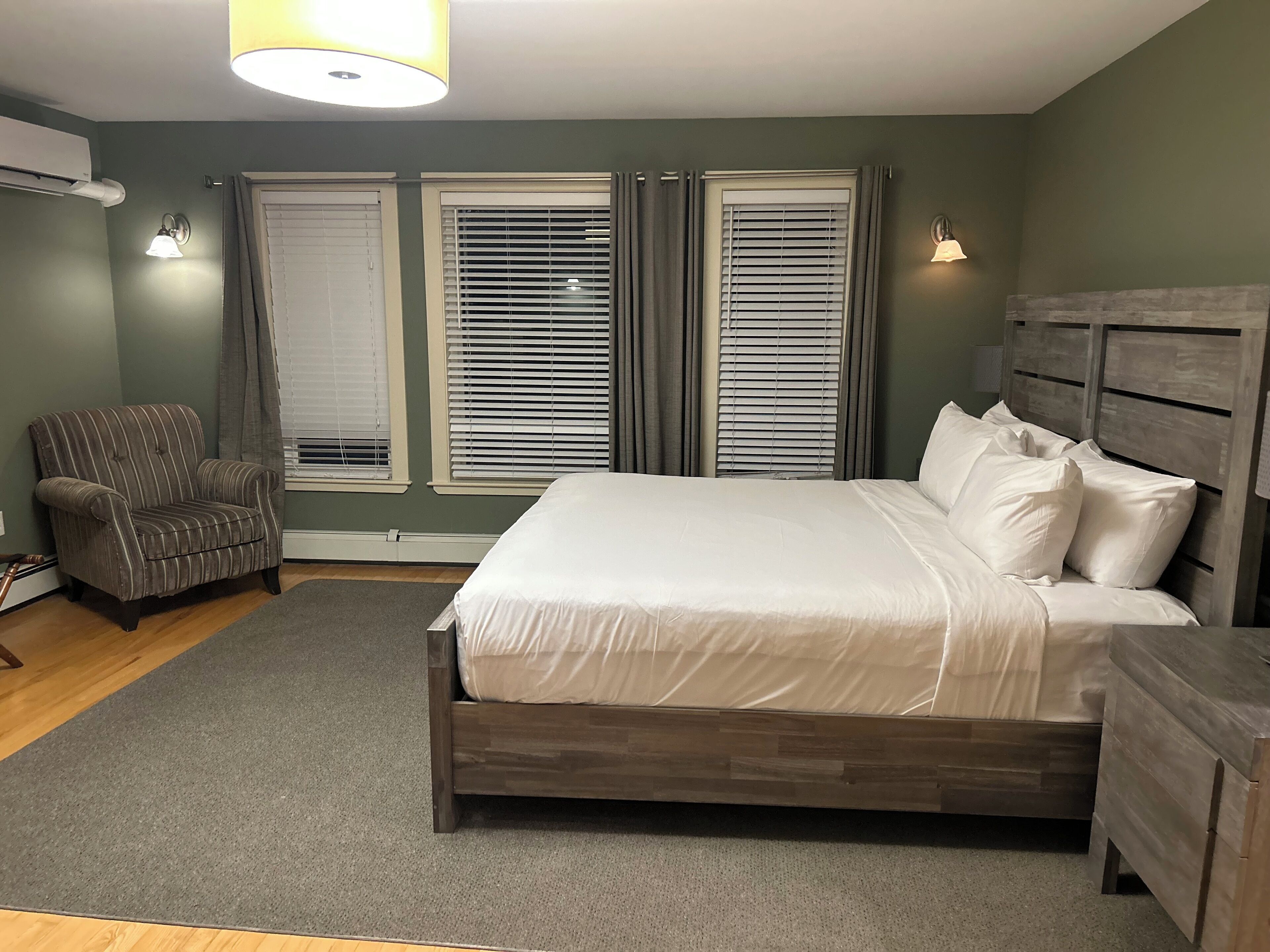 A bedroom with a wooden bed, two windows with blinds, a chair, and a lamp.
