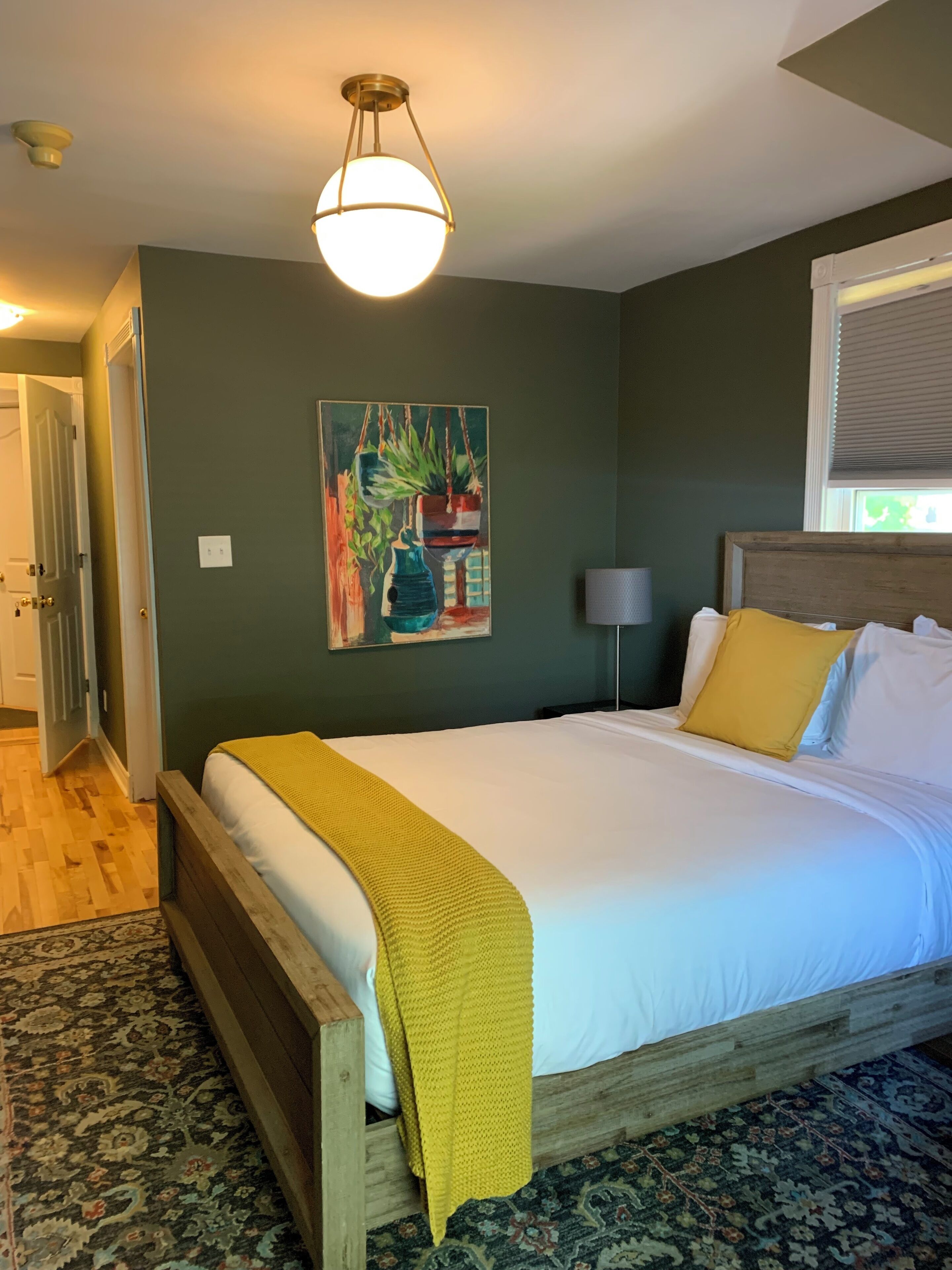 A bedroom with a wooden bed, a yellow throw blanket, a painting on the wall, and a window with blinds.