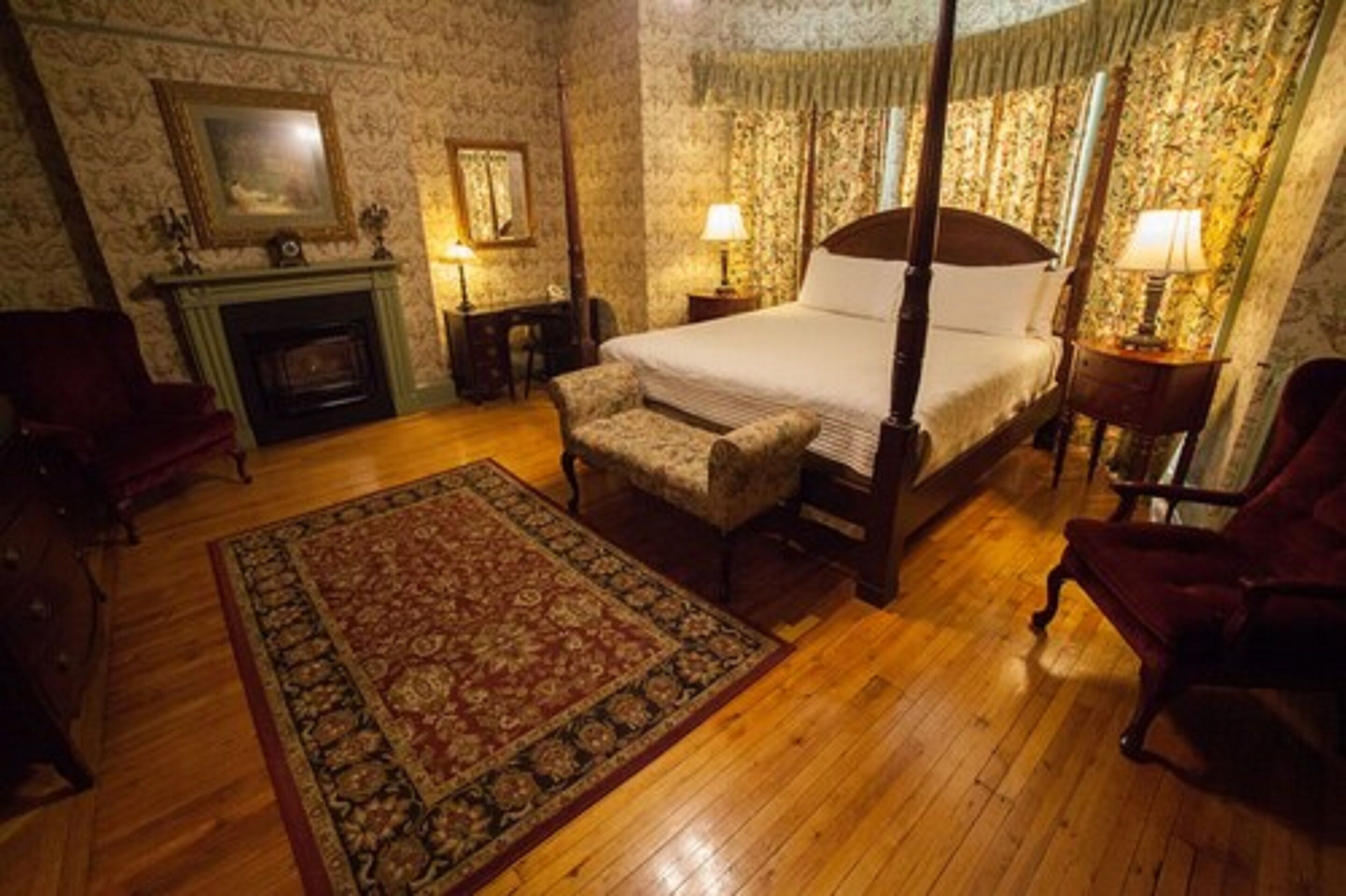 A traditional bedroom with a four-poster bed, a fireplace, a desk, and a red armchair.