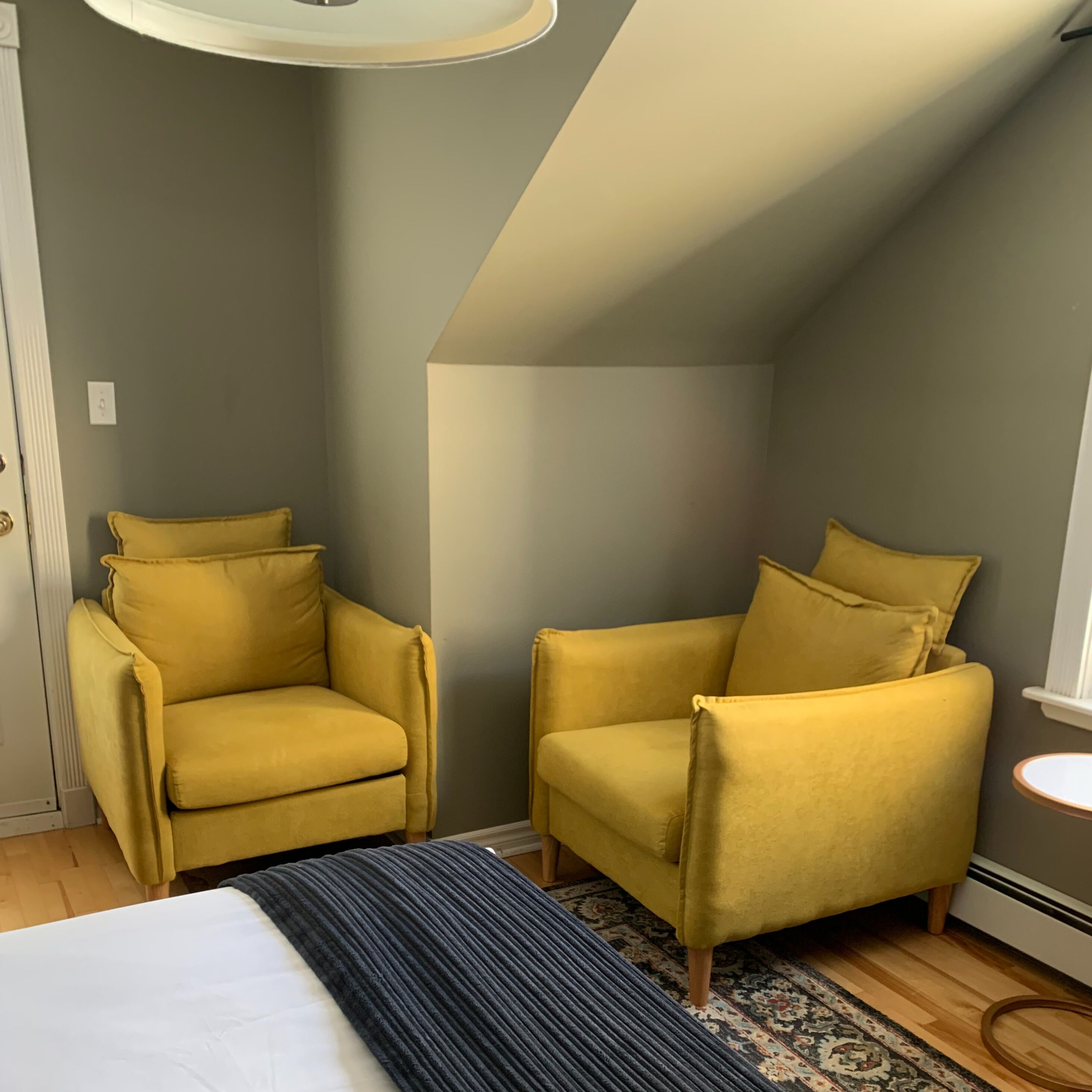 A corner room with two yellow armchairs, a patterned rug, and a bed with a white headboard.