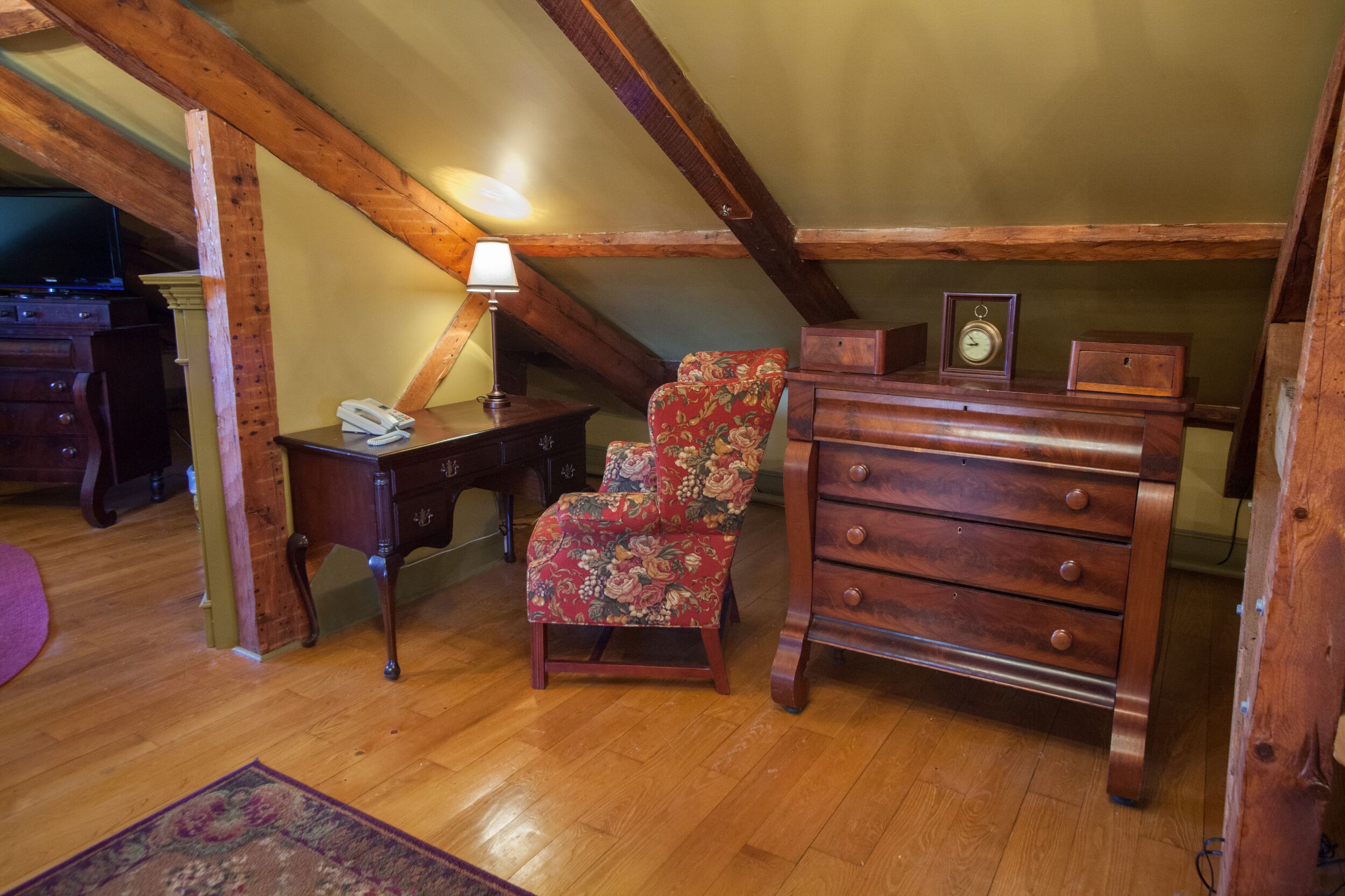 Acogedor dormitorio en áticos con vigas de madera, un sillón floral, un aparador de madera y una lámpara.