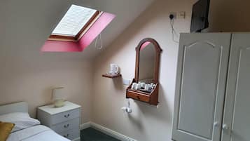 A bedroom with a sloped ceiling, a skylight, a bed with white bedding, a nightstand with a lamp, a dresser, a mirror, and a shelf with toiletries.