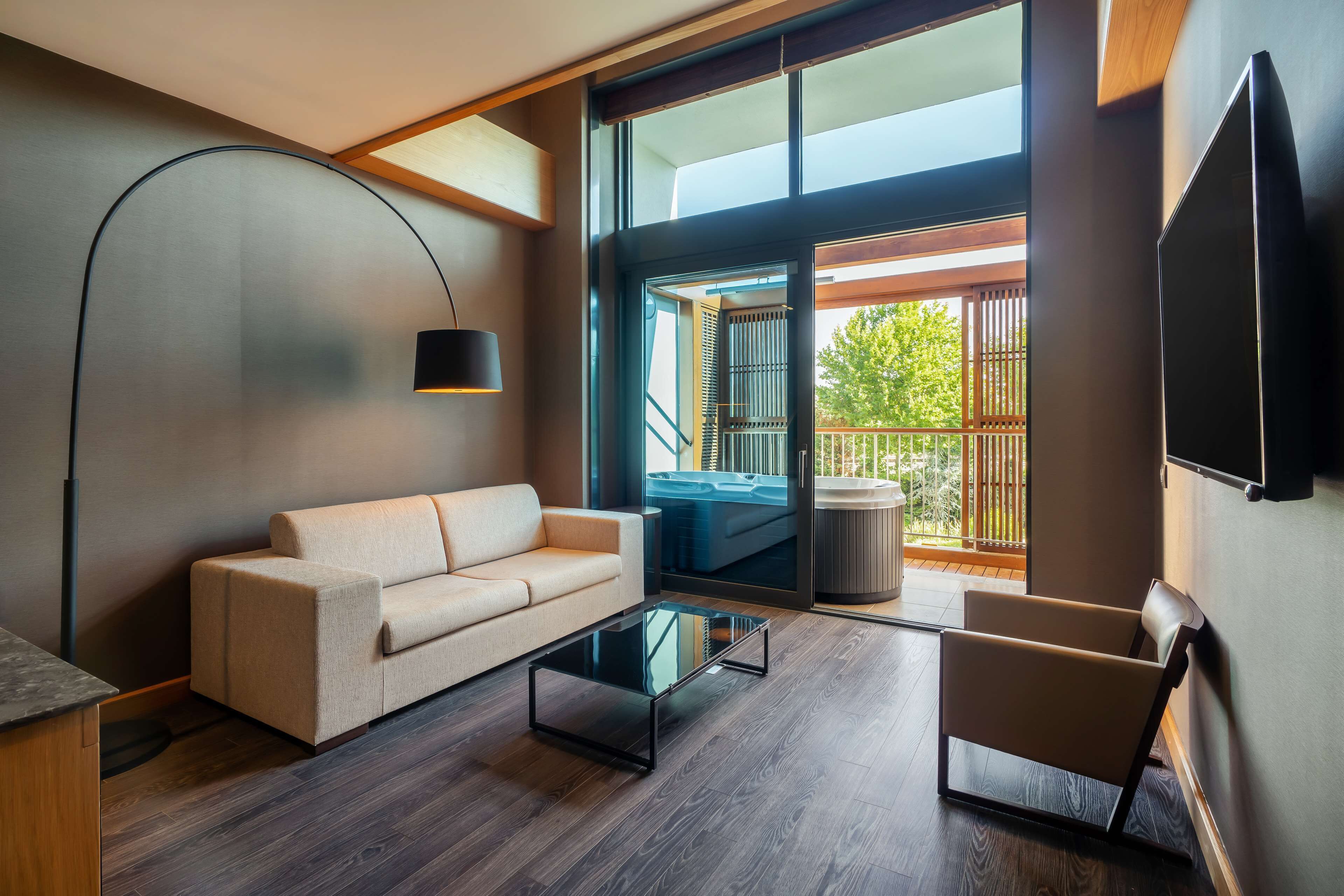 A modern living room with a sofa, a glass coffee table, a chair, a TV, and a large window with a view of a balcony.