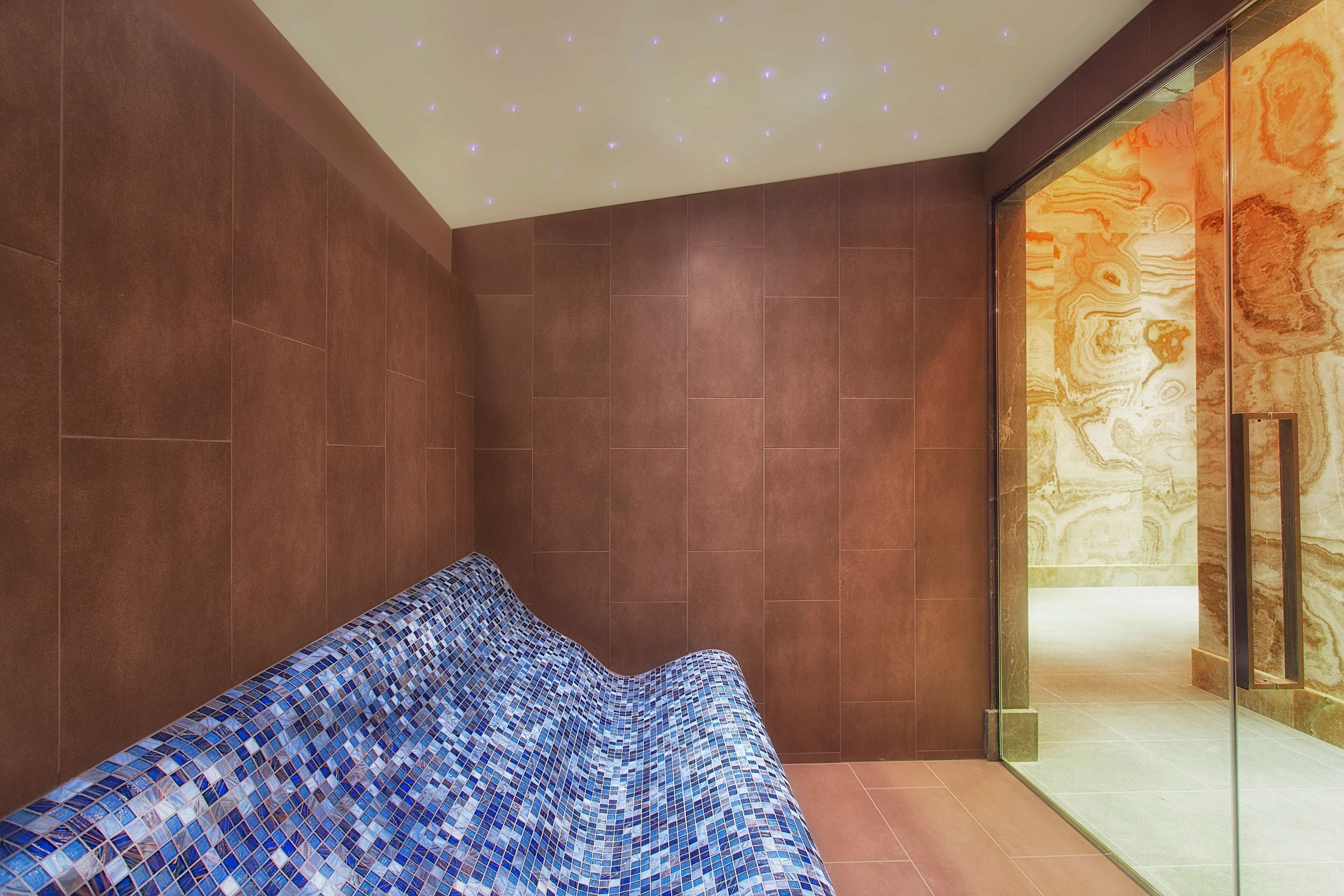 Un espace spa moderne avec un banc orné de mosaïques, des murs de couleur brune et une porte en verre donnant sur une pièce dont l’un des murs est en marbre.