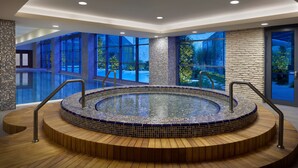Una piscina interior moderna con diseño en mosaico, rodeada de escalones de madera y paredes de cristal que ofrecen vistas al exterior.