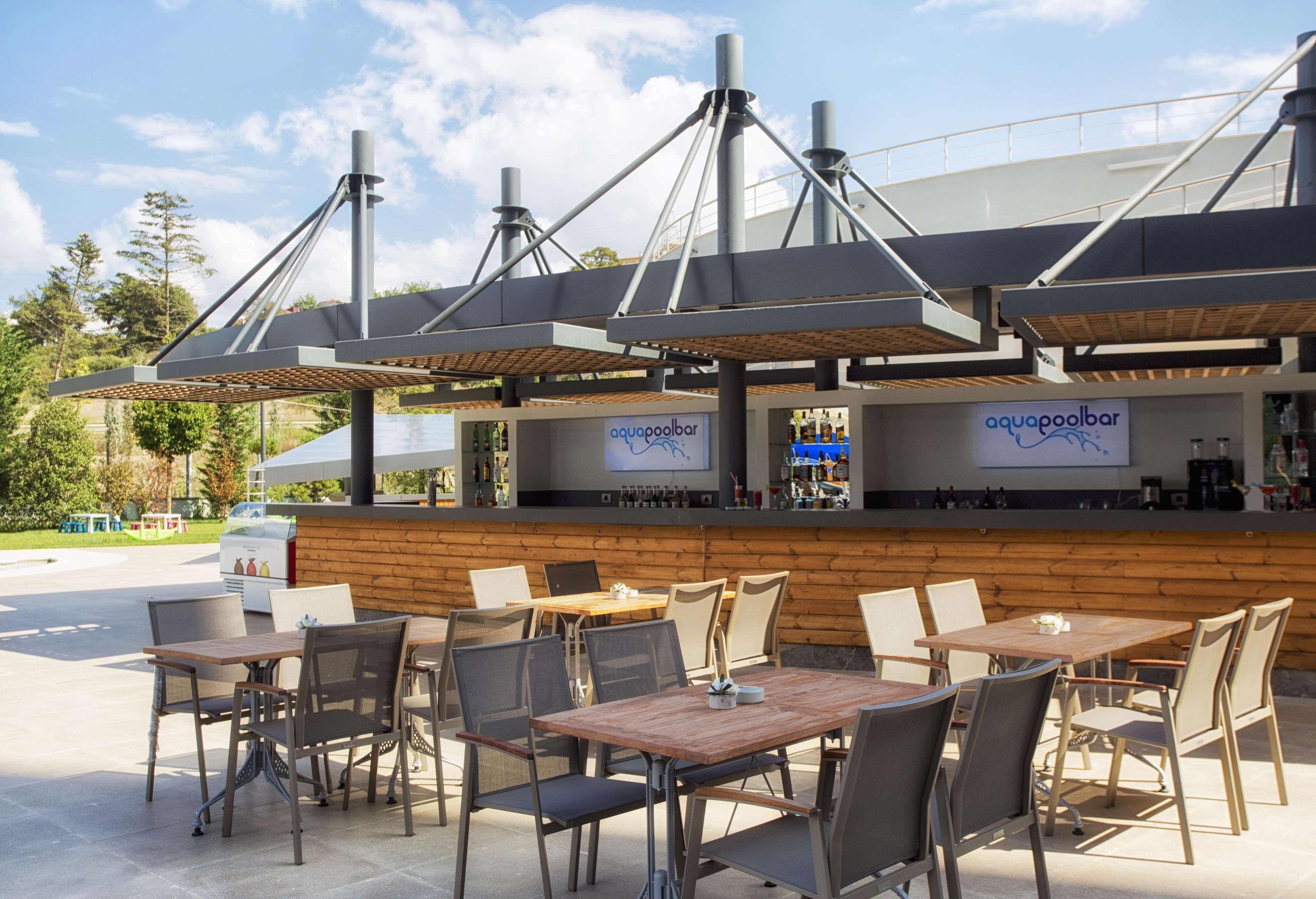 An outdoor seating area with tables and chairs, a bar with a sign, and a view of trees and sky.