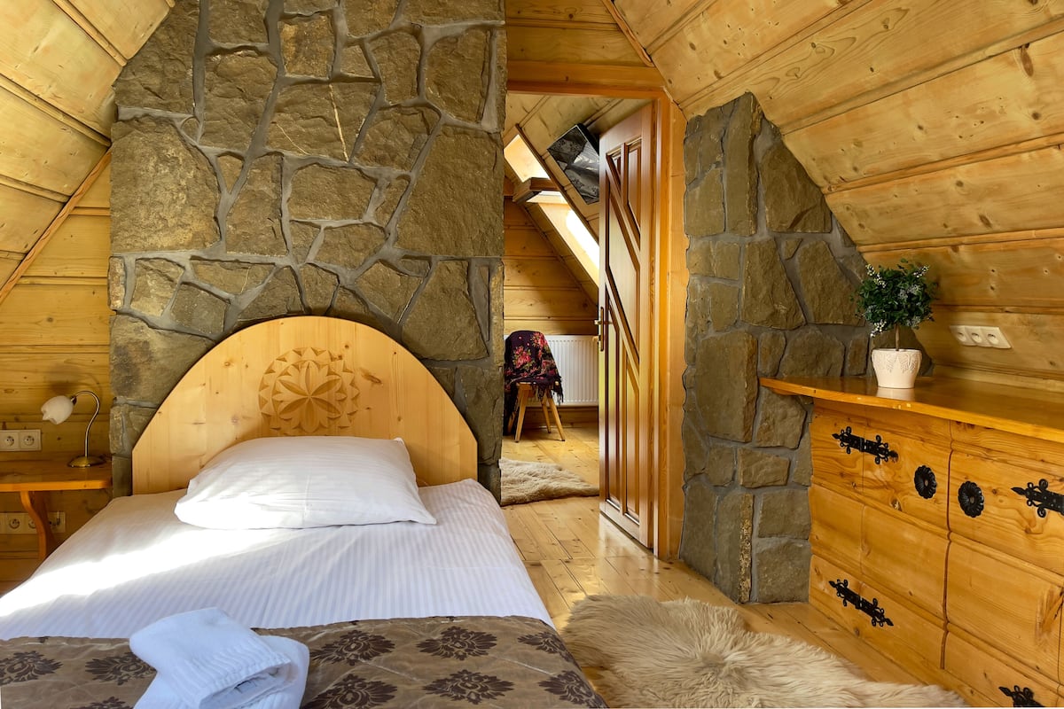 Ein gemütliches Zimmer mit Steinwand, Holzboden, einem Bett mit weißer Bettwäsche, einem Schminktisch mit schwarzen Griffen und einer Topfpflanze.
