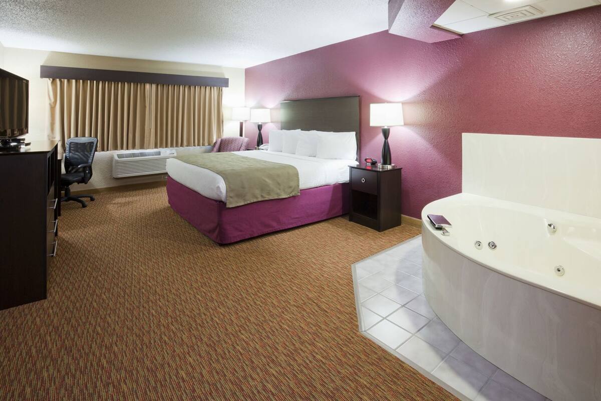 Une chambre d’hôtel comprenant un lit, un bureau avec une chaise, une lampe de bureau, une table de chevet avec une lampe et une baignoire.