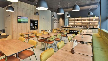 Un café moderno con mesas de madera y sillas amarillas, una zona de cocina con mostrador e iluminación colgante.
