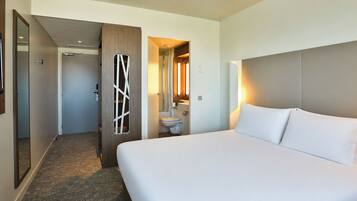 Quarto de hotel com uma cama grande, banheiro com vaso sanitário e pia, e corredor que leva a outro quarto.
