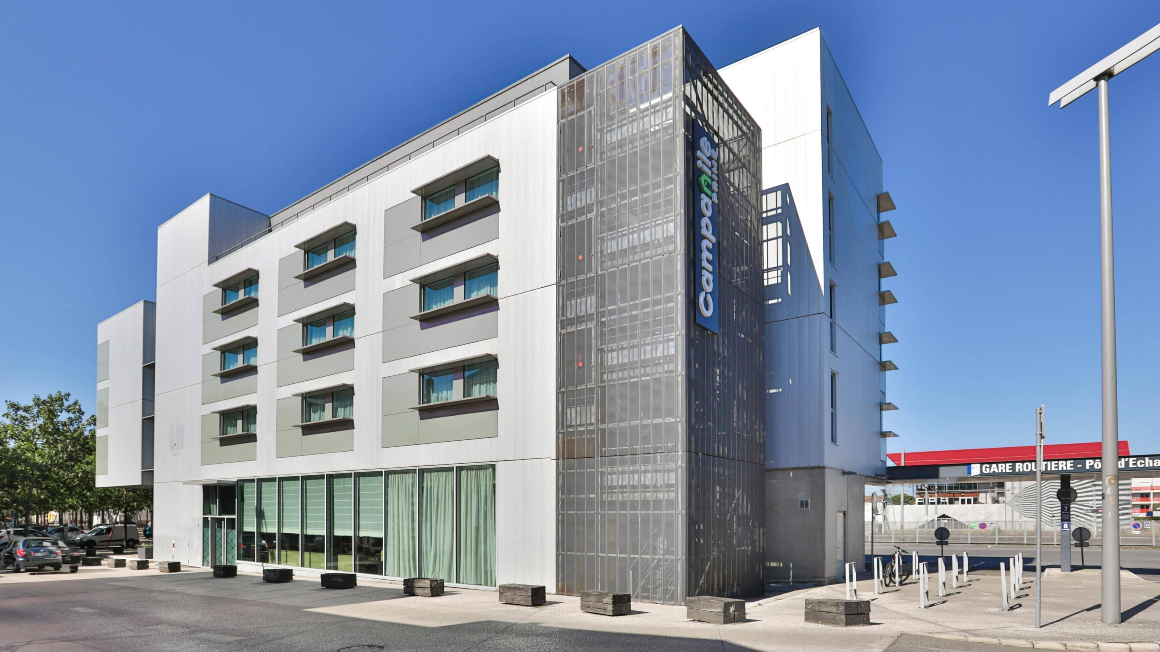 A modern building with a glass facade, multiple floors, and a parking area in front.