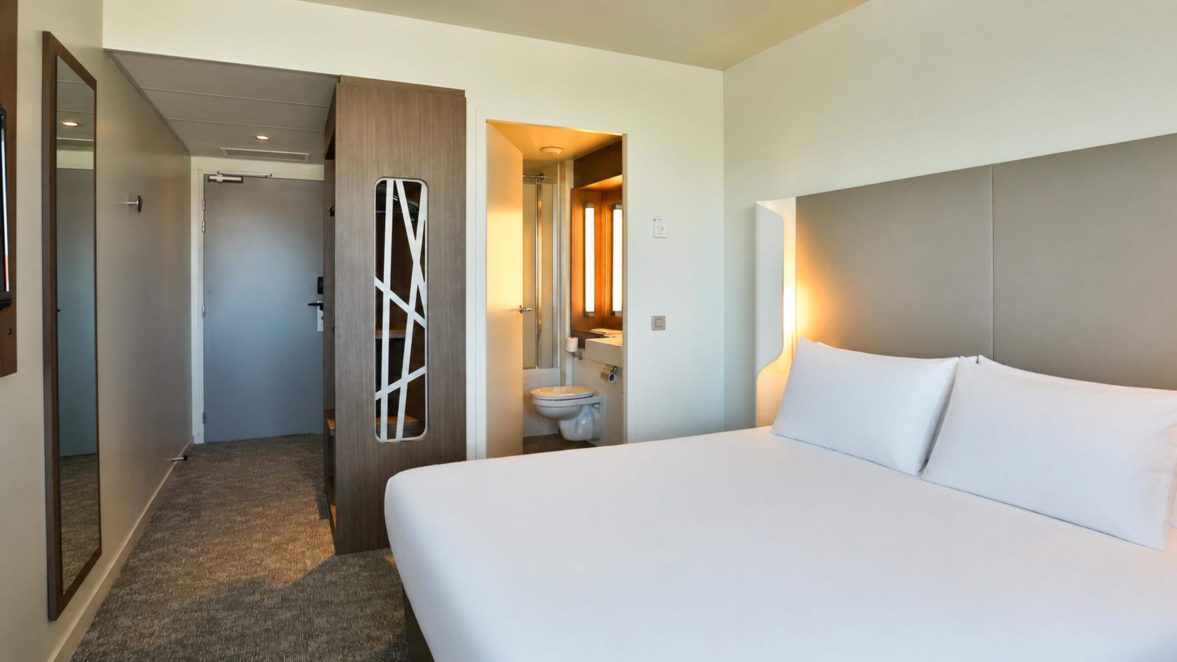 A hotel room with a large bed, a bathroom with a toilet and sink, and a hallway leading to another room.