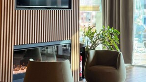 A modern living room with a flat-screen TV mounted on a wooden panel wall, a comfortable sofa, and a large window with curtains. - Campanile PRIME - La Roche Sur Yon Centre Gare (La Roche-sur-Yon)