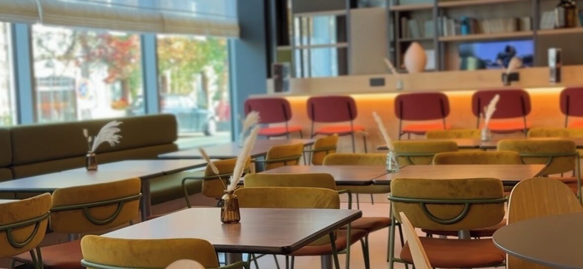 Un café moderne avec des chaises et des tables aux coussins jaunes, un coin bar avec des chaises rouges et de grandes fenêtres donnant sur la rue.