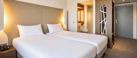 A hotel room with two beds, a bedside table with a phone, a bathroom visible through an open door, and a door with a geometric design.