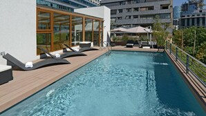 Una piscina al terrat amb gandules, una zona tancada amb vidre i vista als edificis circumdants.