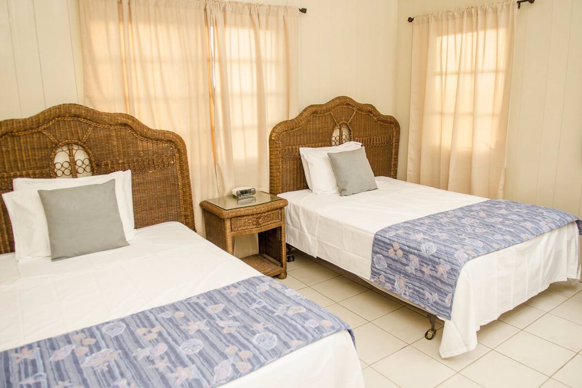 Une chambre d’hôtel avec deux lits simples, une petite table de chevet en bois et une fenêtre avec des rideaux légers.