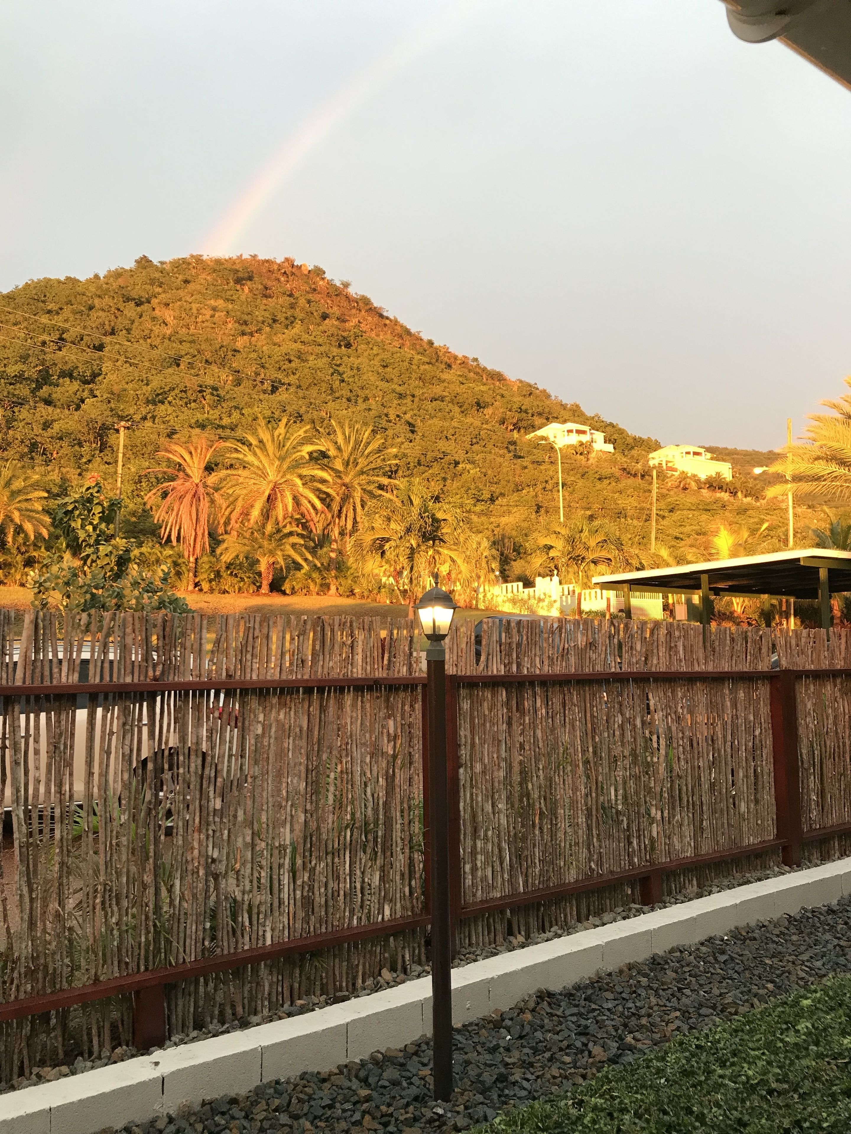 Una cerca de madera, un farol de la calle, una carretera con línea blanca y una colina con árboles y edificios al fondo.