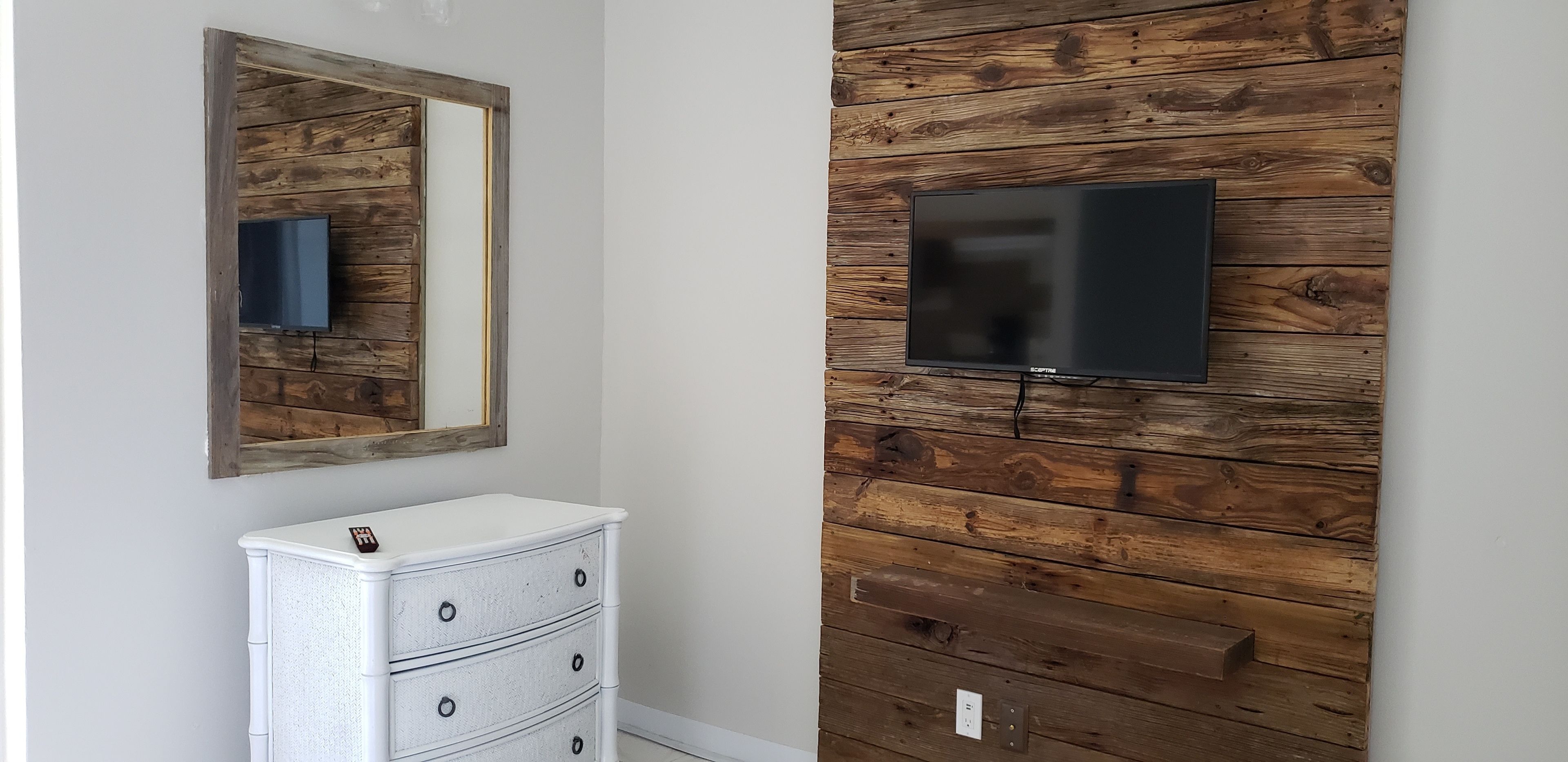 A room with a wooden wall panel, a flat-screen TV mounted on it, and a white dresser with a mirror above it.