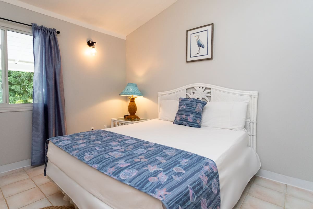 Une chambre à coucher avec un lit, une table de chevet, une lampe, une fenêtre avec des rideaux et un tableau encadré au mur.