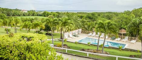 A pool area with lounge chairs and palm trees, surrounded by lush greenery and a view of the ocean.