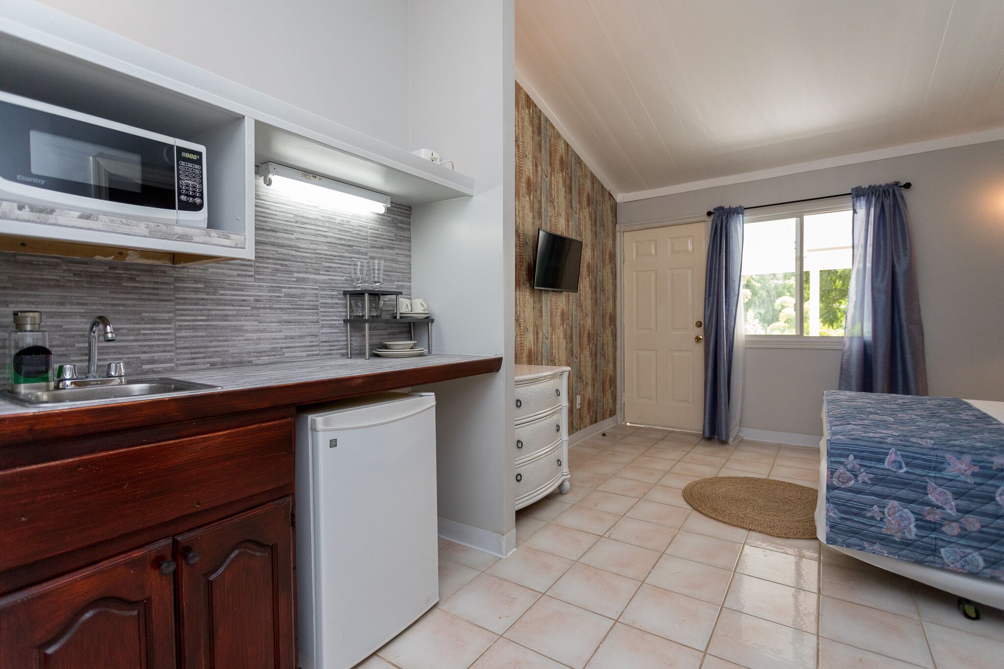 Cocina compacta con microondas, pileta y refrigerador pequeño. Se observa un armario de madera, un buzo blanco y una cama con colcha estampada. Un televisor está montado en la pared, y se ve una ventana con cortinas.